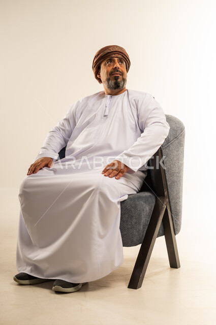 The concept of manhood and dignity, gestures of self-confidence, interest in outward appearance, portrait of an Arab Gulf Omani man wearing a dishdasha and turban sitting on a comfortable sofa looking somewhere with expressions of pleasure, white background
