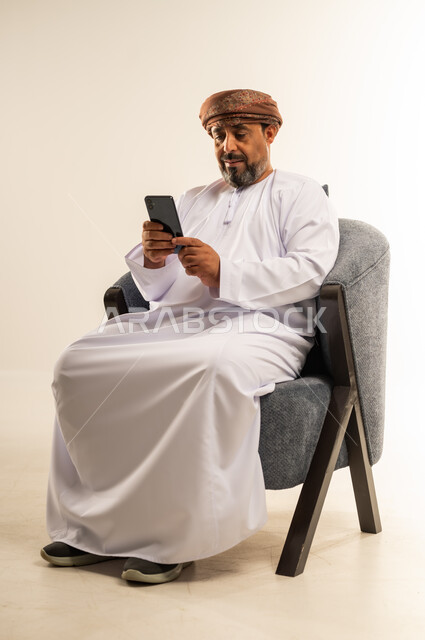 الدردشة مع الاهل والأصدقاء عبر تطبيقات الجوال، تصفح مواقع التواصل الاجتماعي, بورتريه لرجل عربي خليجي عماني يرتدي الدشداشة والعمامة يجلس على كرسي مريح يحمل بيده الهاتف المحمول, استخدام التقنيات الحديثة المتطورة, خلفية بيضاء