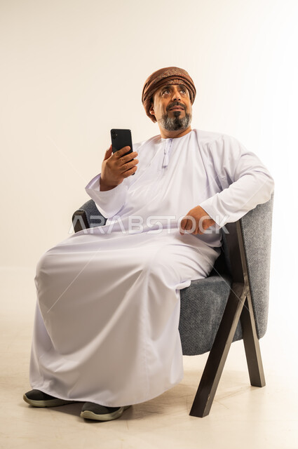 الدردشة مع الاهل والأصدقاء عبر تطبيقات الجوال، تصفح مواقع التواصل الاجتماعي, بورتريه لرجل عربي خليجي عماني يرتدي الدشداشة والعمامة يجلس على كرسي مريح يحمل بيده الهاتف المحمول, استخدام التقنيات الحديثة المتطورة, خلفية بيضاء