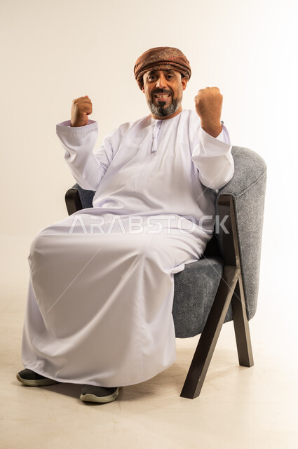 القوة والتحدي والحماس، مفهوم الانجاز وتحقيق الاهداف، بورتريه لرجل عربي خليجي عماني يرتدي الدشداشة والعمامة يجلس على أريكة مريحة يرفع قبضتيه نحو الأعلى بإيماءات التشجيع، تعابير السرور والسعادة، خلفية بيضاء