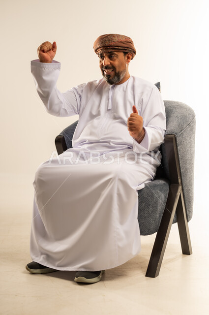 مفهوم الانجاز وتحقيق الاهداف، القوة والتحدي والحماس، بورتريه لرجل عربي خليجي عماني يرتدي الدشداشة والعمامة يجلس على أريكة مريحة يرفع قبضتيه نحو الأعلى بإيماءات التشجيع، تعابير السرور والسعادة، خلفية بيضاء