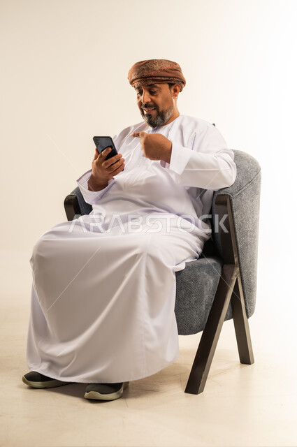 الدردشة مع الاهل والأصدقاء عبر تطبيقات الجوال، تصفح مواقع التواصل الاجتماعي, بورتريه لرجل عربي خليجي عماني يرتدي الدشداشة والعمامة يجلس على كرسي مريح يشير بإصبع السبابة للهاتف المحمول, استخدام التقنيات الحديثة المتطورة, خلفية بيضاء