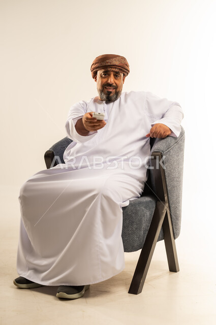 Changing and flipping between channels, the concept of guidance and control, using modern technical devices, a portrait of an Arab Gulf Omani man wearing a dishdasha and turban sitting on a comfortable chair holding a remote control in his hand, watching movies and entertaining programs, white background