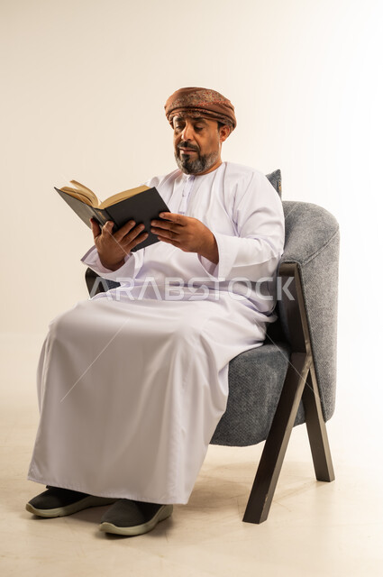 حب المطالعة والقراءة، بورتريه لرجل عربي خليجي عماني  يرتدي الدشداشه والعمامة يجلس على اريكة مريحة يحمل بيده كتاب، تمضية أوقات الفراغ بالتعلم والتثقف، الاهتمام بتصفح الكتب المفيدة، خلفية بيضاء