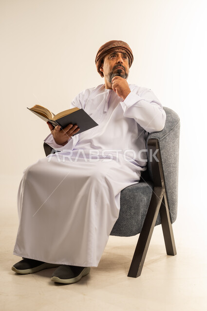 Love of reading and studying, portrait of an Arab Gulf Omani man wearing a dishdasha and turban sitting on a comfortable sofa holding a book in his hand, spending free time learning and educating himself, interest in browsing useful books, white background