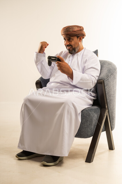 لعب العاب الفيديو، قضاء وقت ممتع في المنزل، انشطة ترفيهية منزلية، استخدام التكنولوجيا الحديثة في الترفيه، بورتريه لرجل عربي خليجي عماني يجلس على الأريكه يمسك بعصا التحكم يرفع يده بايماءات الفوز والنصر، المتعة والتسلية والتفاعل، صورة لكامل الجسم، خلفية بيضاء