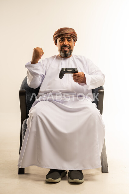 لعب العاب الفيديو، قضاء وقت ممتع في المنزل، انشطة ترفيهية منزلية، استخدام التكنولوجيا الحديثة في الترفيه، بورتريه لرجل عربي خليجي عماني يجلس على الأريكه يمسك بعصا التحكم يرفع يده بايماءات الفوز والنصر، المتعة والتسلية والتفاعل، صورة لكامل الجسم، خلفية بيضاء