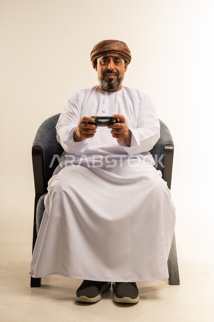 قضاء وقت ممتع في المنزل، انشطة ترفيهية منزلية، استخدام التكنولوجيا الحديثة في الترفيه، بورتريه لرجل عربي خليجي عماني يجلس على الأريكه يمسك بيده عصا التحكم يقوم بلعب العاب الفيديو بإيماءات السعادة والسرور، المتعة والتسلية والتفاعل، صورة لكامل الجسم، خلفية بيضاء