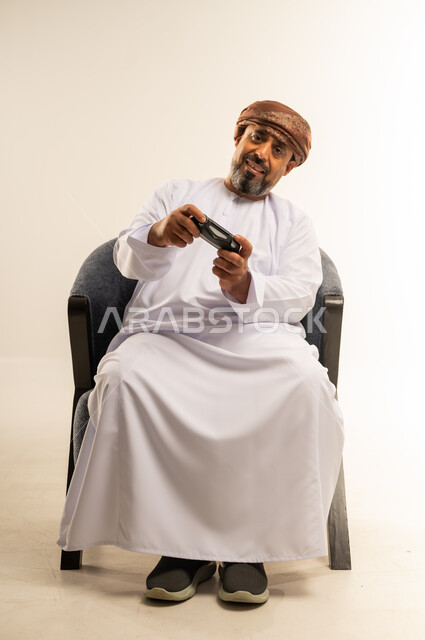 المتعة والتسلية والتفاعل، قضاء وقت ممتع في المنزل، انشطة ترفيهية منزلية، استخدام التكنولوجيا الحديثة في الترفيه، بورتريه لرجل عربي خليجي عماني يجلس على الأريكه يمسك بعصا التحكم يقوم بلعب العاب الفيديو، صورة لكامل الجسم، خلفية بيضاء