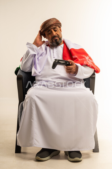 المتعة والتسلية والتفاعل، قضاء وقت ممتع في المنزل، بورتريه لرجل عربي خليجي عماني يرتدي الدشداشة والعمامة يضع على ظهره علم سلطنة عمان يجلس على الأريكه يمسك بيده عصا التحكم يضع يده على رأسه بايماءات الحسرة، لعب العاب الفيديو، صورة لكامل الجسم، خلفية بيضاء