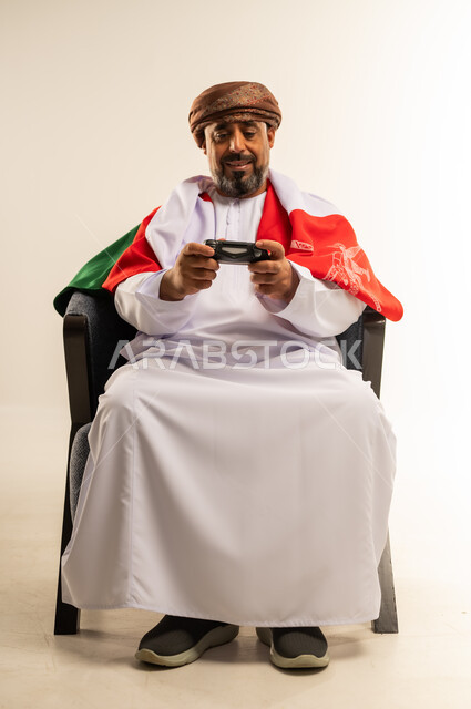 قضاء وقت ممتع في المنزل، استخدام التكنولوجيا الحديثة في الترفيه، بورتريه لرجل عربي خليجي عماني يرتدي الدشداشة والعمامة يضع على ظهره علم سلطنة عمان يجلس على الأريكه يمسك بيده عصا التحكم، لعب العاب الفيديو، المتعة والتسلية والتفاعل، صورة لكامل الجسم، خلفية بيضاء