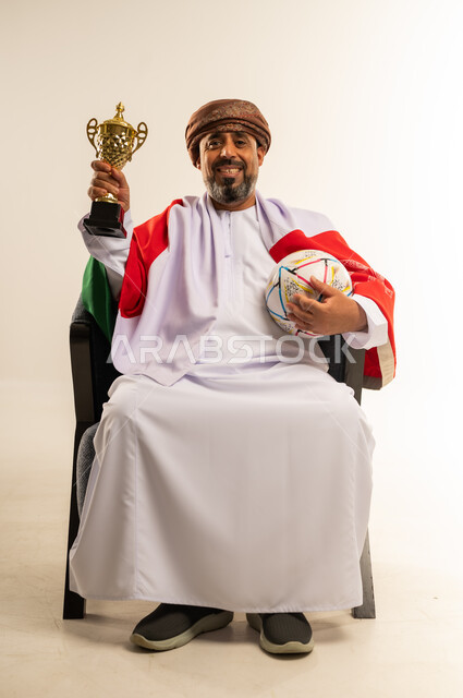 التتويج بكأس البطولة بعد تحقيق الانتصار، تشجيع المنتخب الوطني العماني، بورتريه لرجل عربي خليجي عماني يرتدي الدشداشة العمامة يضع علم سلطنة عمان على كتفيه يجلس على كرسي مريح ويحمل كرة القدم بيده، صورة لكامل الجسم، خلفية بيضاء