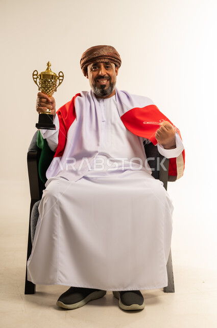 تشجيع المنتخب الوطني العماني، التتويج بكأس البطولة بعد تحقيق الانتصار، بورتريه لرجل عربي خليجي عماني يرتدي الدشداشة العمامة يضع علم سلطنة عمان على كتفيه يجلس على كرسي مريح يرفع يده بايماءات النصر، صورة لكامل الجسم، خلفية بيضاء
