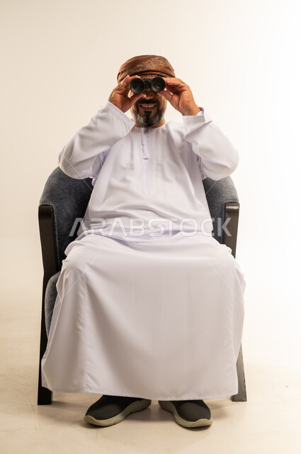 رؤية الاشياء البعيدة وتقريبها، حب الاستكشاف والمغامرة، بورتريه لرجل عربي خليجي عماني يرتدي الدشداشة والعمامة يحمل بيده منظار ثنائي العينين وينظر الى الكاميرا بإيماءات السرور والاستمتاع، صورة الجسم بالطول الكامل، خلفية بيضاء