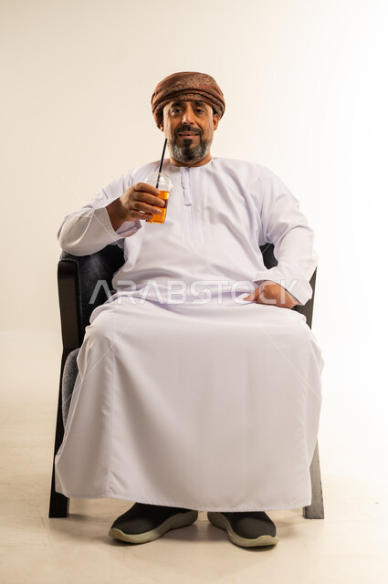 الاستمتاع بتناول المشروبات المفيدة اللذيذة، محاولة التخفيف من حرارة الصيف بالعصائر المنعشة، بورتريه لرجل عربي خليجي عماني يرتدي الدشداشة والعمامة يقوم بشرب العصير الطبيعي البارد، صورة لكامل الجسم، خلفية بيضاء
