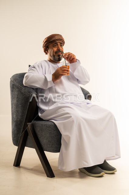 الاستمتاع بتناول المشروبات المفيدة اللذيذة، محاولة التخفيف من حرارة الصيف بالعصائر المنعشة، بورتريه لرجل عربي خليجي عماني يرتدي الدشداشة والعمامة يقوم بشرب العصير الطبيعي البارد، صورة لكامل الجسم، خلفية بيضاء