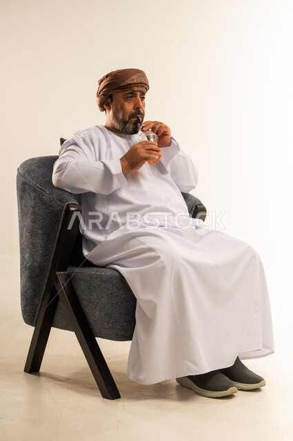 محاولة التخفيف من حرارة الصيف بالعصائر المنعشة، الاستمتاع بتناول المشروبات المفيدة اللذيذة، بورتريه لرجل عربي خليجي عماني يرتدي الدشداشة والعمامة يقوم بشرب العصير الطبيعي البارد، صورة لكامل الجسم، خلفية بيضاء