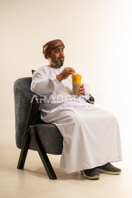 انشطة ترفيهية منزلية، الاستمتاع بتناول الفشار، بورتريه لرجل عربي خليجي عماني يرتدي الدشداشة والعمامة يحمل بيده علبة الذرة، سناكات ومقرمشات لذيذة، صورة لكامل الجسم، خلفيه بيضاء