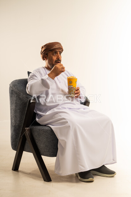 الاستمتاع بتناول الفشار، انشطة ترفيهية منزلية، بورتريه لرجل عربي خليجي عماني يرتدي الدشداشة والعمامة يحمل بيده علبة الذرة، سناكات ومقرمشات لذيذة، صورة لكامل الجسم، خلفيه بيضاء