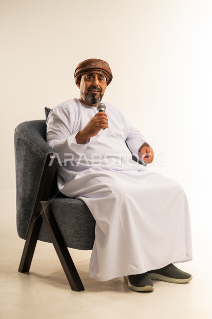 الدورات التدريبية و ورشات العمل، إلقاء المحاضرات وإقامة الندوات، تبادل الحوار والاستفادة من خبرات كبار السن، بورتريه لرجل عربي خليجي عماني يرتدي الدشداشة والعمامة يجلس على أريكة مريحة يحمل بيده مكبر الصوت، صورة الجسم بالطول الكامل، خلفية بيضاء