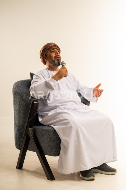 إلقاء المحاضرات وإقامة الندوات، الدورات التدريبية و ورشات العمل، تبادل الحوار والاستفادة من خبرات كبار السن، بورتريه لرجل عربي خليجي عماني يرتدي الدشداشة والعمامة يجلس على أريكة مريحة يحمل بيده مكبر الصوت، صورة الجسم بالطول الكامل، خلفية بيضاء