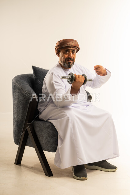 الاشارة بإصبع السبابة إلى الدامبلز، بناء عضلات الجسم وتقويتها، رفع الأوزان الحديدية الثقيلة، بورتريه لرجل عربي خليجي عماني يرتدي الدشداشة والعمامة يجلس على الاريكة يحمل بيده الأثقال، نشاط بدني للمحافظة على اللياقة الجسدية، الطول الكامل للجسم، خلفية بيضاء