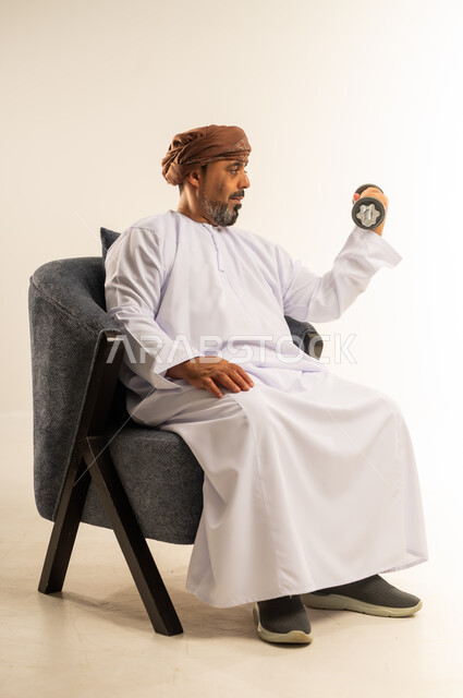 رفع الأوزان الحديدية الثقيلة، بناء عضلات الجسم وتقويتها، بورتريه لرجل عربي خليجي عماني يرتدي الدشداشة والعمامة يجلس على الاريكة يحمل بيده الأثقال، نشاط بدني للمحافظة على اللياقة الجسدية، الطول الكامل للجسم، خلفية بيضاء