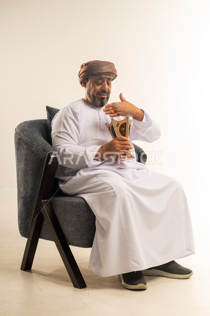 مفهوم التطيب بالبخور, استخدام خشب العود بالأعياد والمناسبات, العادات والتقاليد العمانية, بورتريه لرجل عربي خليجي عماني يرتدي الدشداشة والعمامة يجلس على كرسي مريح يقوم بالتعطر من المبخرة, الاهتمام بالمظهر الخارجي, خلفية بيضاء