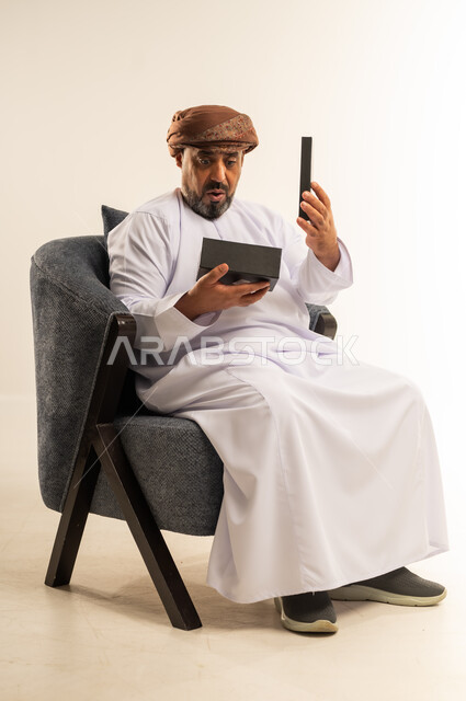 مفهوم الشكر والامتنان، تقديم الهدايا بالاعياد والمناسبات الخاصة، التعبير عن الحب والتقدير والاحترام، بورتريه لرجل عربي خليجي عماني يرتدي الدشداشة والعمامة يجلس على أريكة مريحة يحمل بيديه صندوق هدية, ايماءات المفاجئة والصدمة، خلفية بيضاء