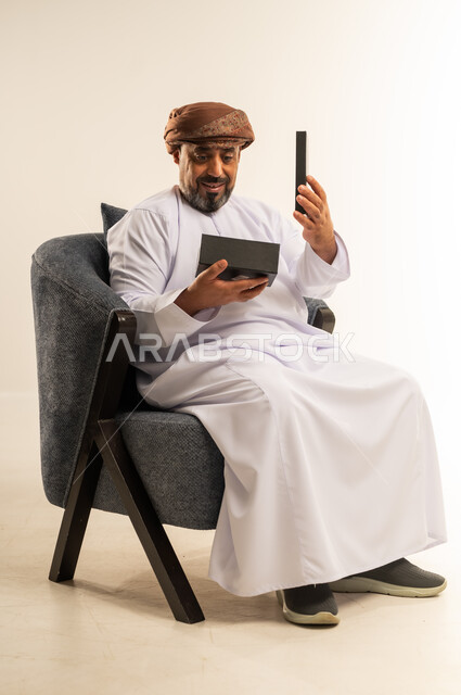 تقديم الهدايا بالاعياد والمناسبات الخاصة، مفهوم الشكر والامتنان، التعبير عن الحب والتقدير والاحترام، بورتريه لرجل عربي خليجي عماني يرتدي الدشداشة والعمامة يجلس على أريكة مريحة يحمل بيديه صندوق هدية, ايماءات المفاجئة والصدمة، خلفية بيضاء