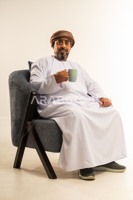Enjoying hot drinks, eating warm winter herbs, spending free time sipping favorite drink, portrait of a smiling Omani Gulf Arab man wearing a dishdasha and turban sitting on a comfortable sofa drinking coffee with gestures of happiness and pleasure, white background