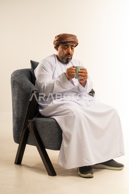 Eating warm winter herbs, enjoying hot drinks, spending free time sipping your favorite drink, portrait of a smiling Omani Gulf Arab man wearing a dishdasha and turban sitting on a comfortable sofa drinking coffee with gestures of happiness and pleasure, white background