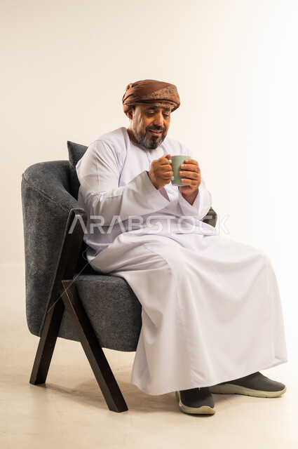 قضاء وقت الفراغ باحتساء المشروب المفضل, تناول الأعشاب الشتوية الدافئة، الاستمتاع بالمشروبات الساخنة، بورتريه لرجل عربي خليجي عماني مبتسم يرتدي الدشداشة والعمامة يجلس على اريكة مريحة يقوم بشرب القهوة بإيماءات السعادة والسرور، خلفية بيضاء