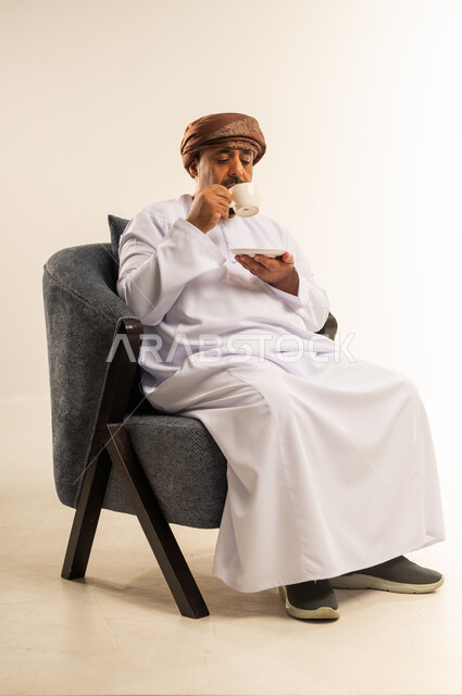 تناول الأعشاب الشتوية الدافئة، الاستمتاع بالمشروبات الساخنة، قضاء وقت الفراغ باحتساء المشروب المفضل, بورتريه لرجل عربي خليجي عماني مبتسم يرتدي الدشداشة والعمامة يجلس على اريكة مريحة يقوم بشرب القهوة بإيماءات السعادة والسرور، خلفية بيضاء