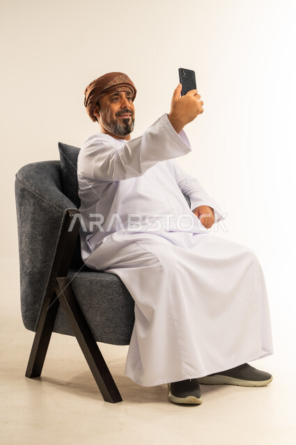 التقاط صور السلفي التذكارية، الدردشة والتواصل مع الاصدقاء بإيماءات السعادة والفرح، بورتريه لرجل عربي خليجي عماني يرتدي الدشداشة والعمامة يمسك بيده الهاتف المحمول يقوم بإجراء مكالمة فيديو، خلفية بيضاء