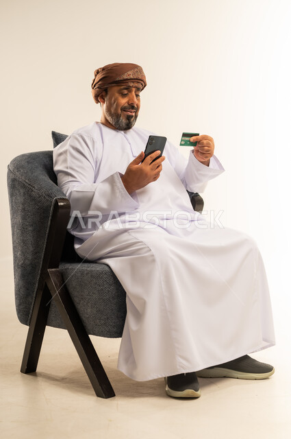 استخدام الأجهزة التقنية الحديثة، إمكانية تسديد الفواتير الانترنت, سهولة الشراء والدفع عن طريق بطاقة الإئتمان البنكية، بورتريه لرجل عربي خليجي عماني يرتدي الدشداشة والعمامة يجلس على كرسي مريح يمسك بيده كرت الصراف و الهاتف المحمول، خلفية بيضاء