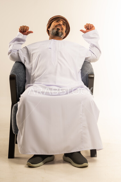 Locating something, hand gestures indicating this place (here), portrait of a smiling Omani Gulf Arab man wearing a dishdasha and turban sitting on a comfortable sofa pointing his index fingers backwards in gestures of pleasure and happiness, white background