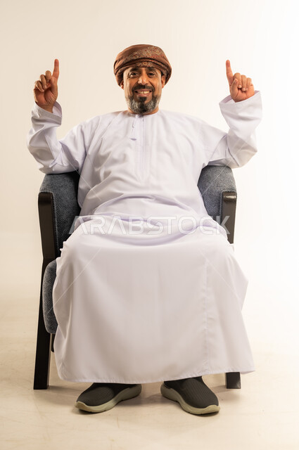 حركات يدين تدل على هذا المكان (هنا)، تحديد موقع شيء ما، بورتريه لرجل عربي خليجي عماني مبتسم يرتدي الدشداشة والعمامة يجلس على أريكة مريحة يشير بأصبعي السبابة نحو الاعلى بايماءات السرور والسعادة، خلفية بيضاء