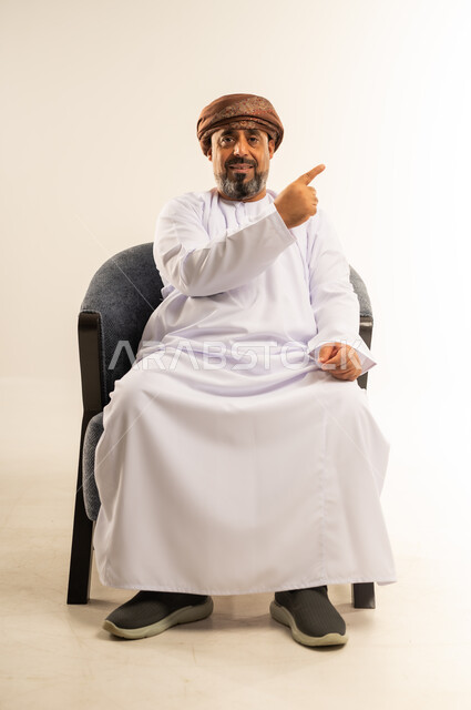 اشارات يدين تدل على هذا المكان (هنا)، تحديد موقع شيء ما، بورتريه لرجل عربي خليجي عماني مبتسم يرتدي الدشداشة والعمامة يجلس على أريكة مريحة يشير بأصبعي السبابة نحو اليسار بايماءات السرور والسعادة، خلفية بيضاء
