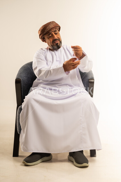 Concern about appearance, expressions of self-confidence, concept of masculinity and dignity, portrait of an Arab Gulf Omani man wearing a dishdasha and turban sitting on a comfortable chair adjusting his sleeve with gestures of pleasure, white background