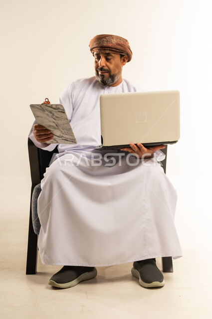 التدقيق في قراءة الملاحظات، مراجعة ومتابعة الخطط والمشاريع المكتبية، اعداد التقارير الخاصة بالعمل، بورتريه لرجل عربي خليجي عماني يرتدي الدشداشة والعمامة يحمل الحاسوب المحمول وحافظة الاوراق، خلفية بيضاء