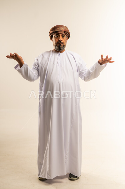 Looking at the camera with gestures of surprise, uncertainty and inability to make a decision, hesitation about a certain matter, portrait of an Arab Gulf Omani man wearing a dishdasha and turban raising his hands up with an expression of confusion, full body image, white background