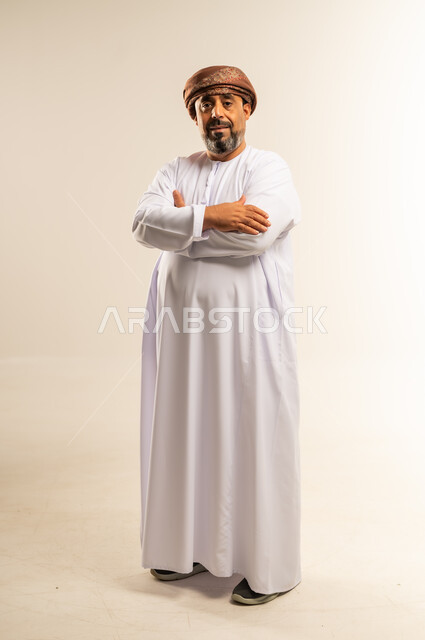 تعابير الثقة بالنفس، الاهتمام بالمظهر الخارجي، مفهوم الرجولة والوقار، بورتريه لرجل عربي خليجي عماني يرتدي الدشداشة والعمامة يقف بايدي متقاطعة ينظر إلى الكاميرا بايماءات التركيز، صورة الجسم بالطول الكامل، خلفية بيضاء
