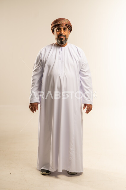 تعابير الثقة بالنفس، مفهوم الرجولة والوقار، الاهتمام بالمظهر الخارجي، بورتريه لرجل عربي خليجي عماني يرتدي الدشداشة والعمامة يقف باستقامة ينظر الى الكاميرا ما بايماءات السرور، صورة الجسم بالطول الكامل، خلفية بيضاء