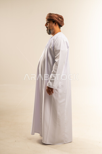 Gestures of self-confidence, concept of masculinity and dignity, interest in outward appearance, side portrait of an Arab Gulf Omani man wearing a dishdasha and turban standing straight looking at the place with expressions of pleasure, full-length body image, white background