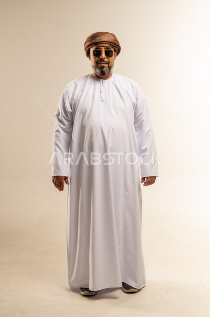 الاهتمام بالمظهر الخارجي، مفهوم الرجولة والوقار، تعابير الثقة بالنفس، بورتريه لرجل عربي خليجي عماني يرتدي الدشداشة والعمامة يضع النظارة الشمسية يقف باستقامة ينظر الى الكاميرا بتعابيرالسرور، صورة الجسم بالطول الكامل، خلفية بيضاء