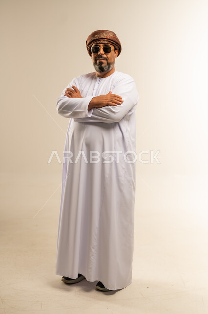 تعابير الثقة بالنفس، الاهتمام بالمظهر الخارجي، مفهوم الرجولة والوقار، بورتريه لرجل عربي خليجي عماني يرتدي الدشداشة والعمامة يضع النظارة الشمسية يقف بايدي متقاطعة ينظر إلى الكاميرا بايماءات التركيز، صورة الجسم بالطول الكامل، خلفية بيضاء