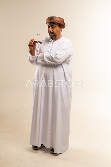 الاهتمام بالمظهر الخارجي، مفهوم الرجولة والوقار، تعابير الثقة بالنفس، بورتريه لرجل عربي خليجي عماني يرتدي الدشداشة والعمامة يضع النظارة الشمسية يقف بايدي متقاطعة، صورة الجسم بالطول الكامل، خلفية بيضاء
