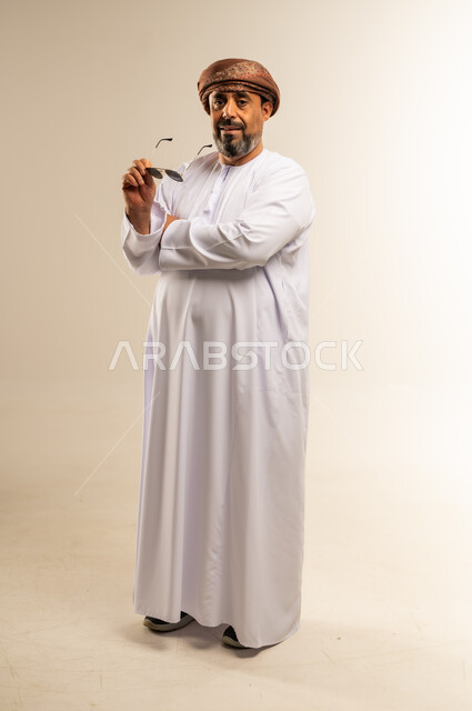 The concept of masculinity and dignity, expressions of self-confidence, interest in external appearance, portrait of an Arab Gulf Omani man wearing a dishdasha and turban, wearing sunglasses, standing with crossed hands looking at the camera with focus gestures, full-length body image, white background
