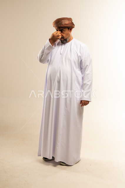 الشعور بالإحباط والخيبة لخسارة شيء ما، إغماض العينين بإيماءات الحزن والرغبة بالبكاء، بورتريه لرجل عربي خليجي عماني يرتدي الدشداشة والعمامة يضع يده على رأسه بتعابير الارهاق، صورة لكمل الجسم، خلفية بيضاء